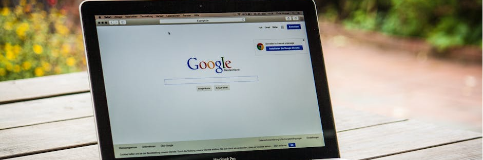 A MacBook Pro displaying Google Search on a wooden table outdoors, next to a smartphone.